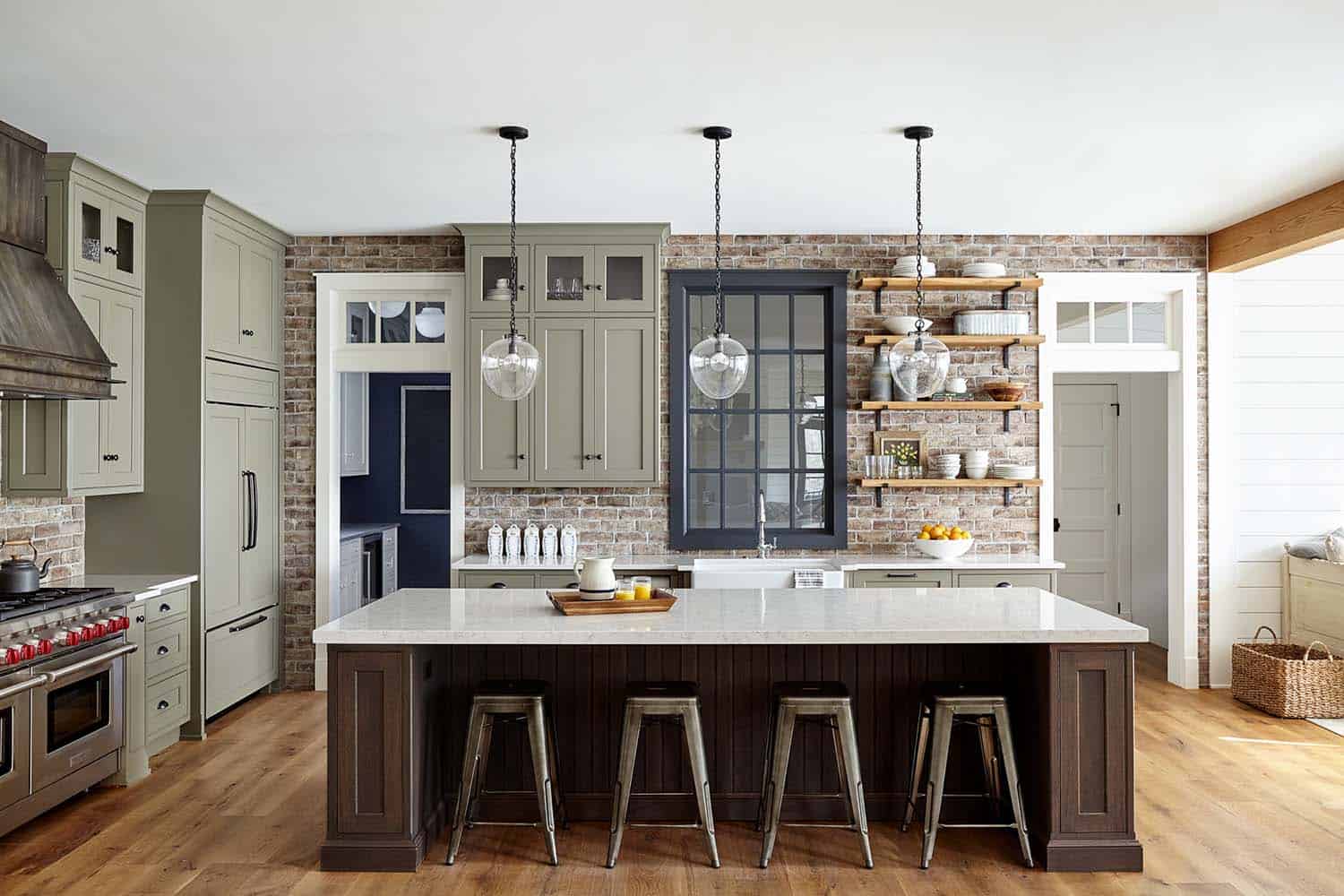 Warm Pennsylvania farmhouse kitchen with sage-green shaker cabinets, exposed brick wall, large island, and natural daylight.
